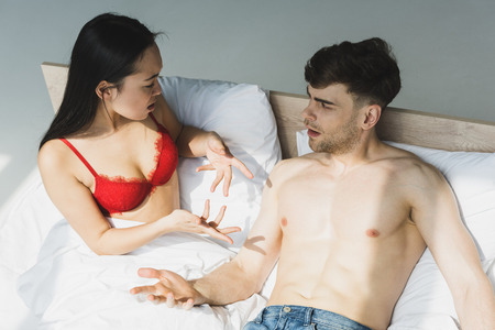 multicultural couple quarreling while lying on white bedding in sunlight