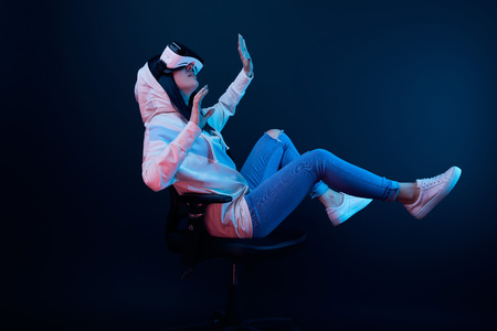 Surprised young woman gesturing and using virtual reality headset while sitting on chair on blue...