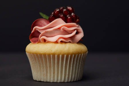 Sweet cupcake with cream and garnet on grey surface isolated on black backgroundの写真素材