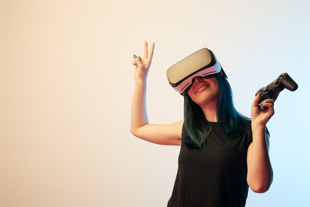 Cheerful brunette woman holding joystick while showing peace sign and wearing virtual reality...