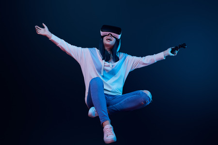 Happy woman in virtual reality headset levitating and holding joystick on blue background