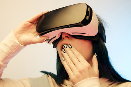 Low angle view of surprised woman covering mouth while wearing virtual reality headset on beige and...