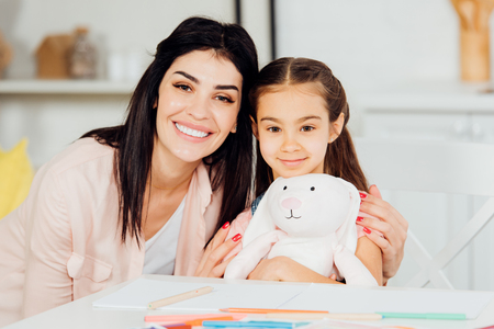 cheerful daughter holding soft toy near brunette mother and looking at cameraの写真素材