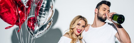 Panoramic shot of cheerful woman holding balloons near man drinking champagne from bottle on white...