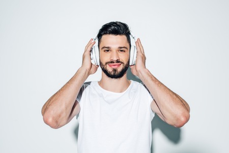 Cheerful man touching headphones while listening music and smiling on white backgroundの写真素材