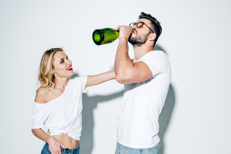 Cheerful blonde girl looking at handsome man in glasses drinking champagne from bottle  on white backgroundの写真素材