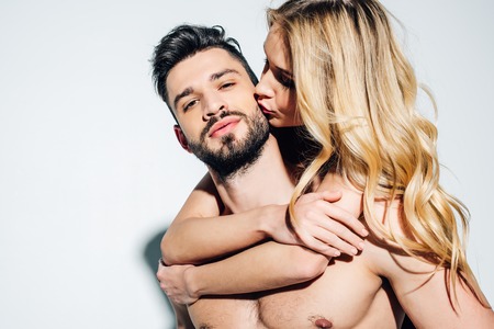 Blonde woman kissing cheek of sexy shirtless man on white background