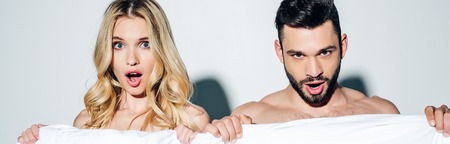 Panoramic shot of surprised blonde woman and handsome man looking at camera while holding blanket on...