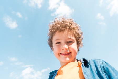 Low angle view of smiling curly kid under blue sky background