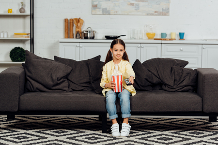 Cute kid holding remote controller while sitting on sofa with bucket of popcornの写真素材