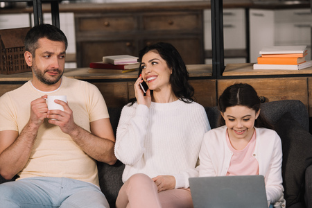happy woman talking on smartphone near daughter with laptop and husband with cup