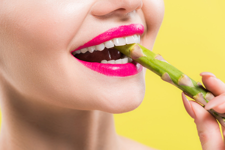 cropped view of cheerful woman holding green and tasty asparagus in mouth isolated on yellowの写真素材