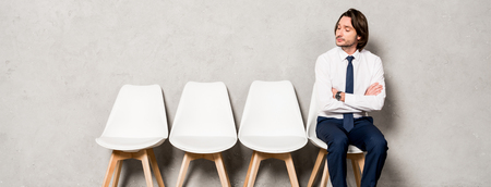 panoramic shot of handsome man sitting on chair with crossed arms and waiting job interviewの写真素材