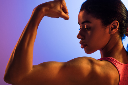 beautiful athletic african american girl demonstrating biceps on purple and blue gradient backgroundの写真素材