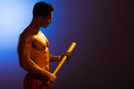 sportive shirtless mixed race man holding baseball bat on blue and dark purple gradient backgroundの写真素材