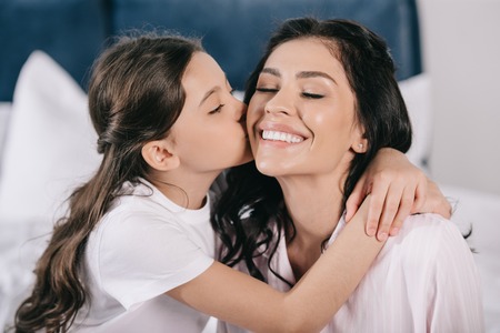 cheerful kid kissing cheek of happy mother and looking at cameraの写真素材
