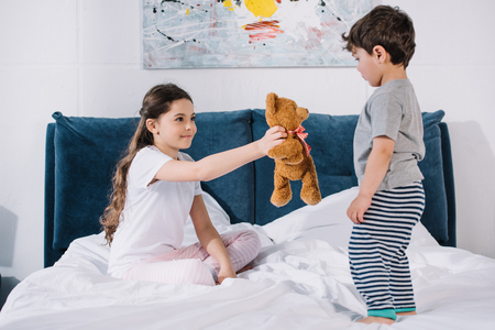 happy kid smiling while holding teddy bear near toddler brother in bedroomの写真素材