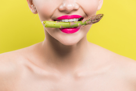 cropped view of cheerful naked woman holding green and tasty asparagus in mouth isolated on yellowの写真素材