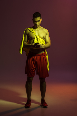 handsome mixed raxe man in red shorts and sneakers using smartphone on dark backgroundの写真素材
