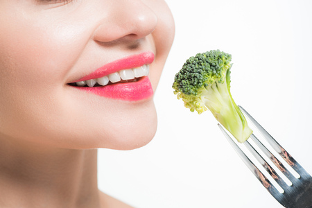 cropped view of happy woman holding silver fork with broccoli near pink lips isolated on whiteの写真素材