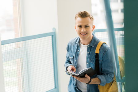 smiling student in denim shirt with yellow backpack holding notebook in universityの写真素材