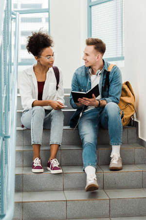 two multiethinc students with notebook sitting on stairs in universityの写真素材