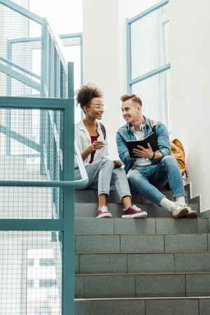smiling multiethnic students with notebook sitting on stairs in collegeの写真素材