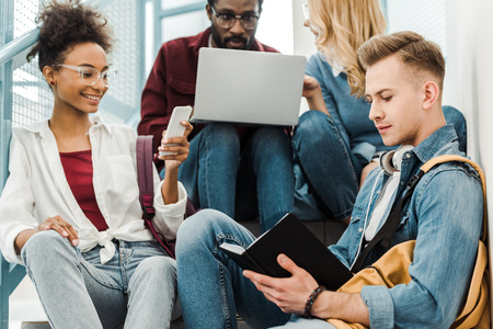 four smiling multiethnic students using laptop and smartphone in collegeの写真素材