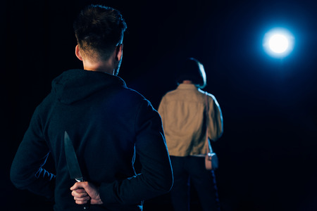 back view view of murderer hiding knife and looking at victim on blackの写真素材