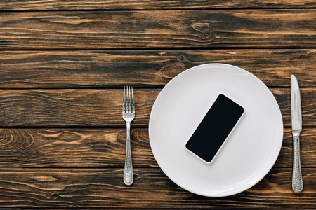 top view of smartphone with blank screen on white plate near knife and fork on brown wooden surfaceの写真素材