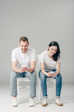 young smiling man and woman sitting on chairs and using smartphones on grey backgroundの写真素材