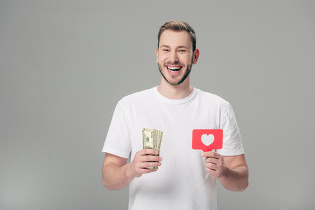 happy handsome man in white t-shirt holding dollar banknotes and red paper cut card with heart symbol isolated on greyの写真素材