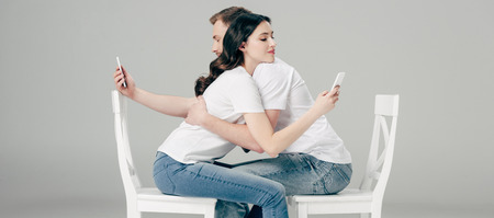 panoramic shot of young man and woman hugging while sitting on white chairs and using smartphones on grey backgroundの写真素材