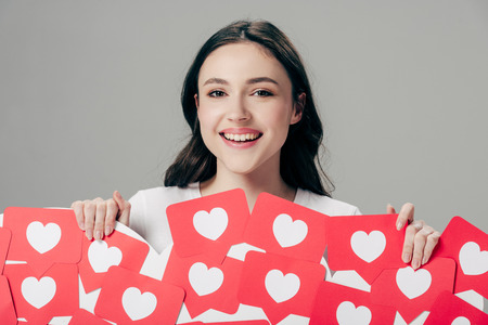cheerful young girl holding red paper cut cards with hearts symbol and looking at camera isolated on greyの写真素材