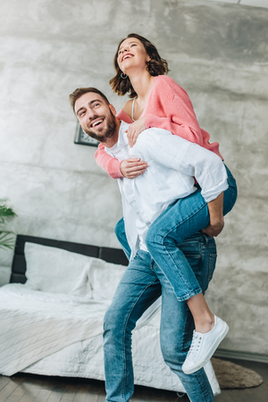 Low angle view of happy bearded man piggybacking brunette woman in bedroomの写真素材