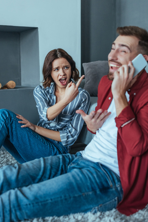 Selective focus of emotional woman showing middle finger while looking at happy man talking on smartphoneの写真素材