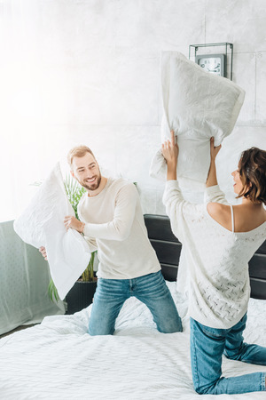 Happy bearded man having pillow fight with cheerful woman on bedの写真素材