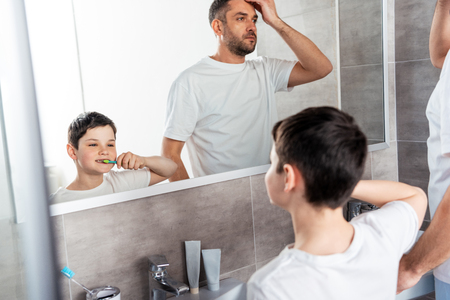 Son brushing teeth while father adjusting hairstyle in bathroomの写真素材