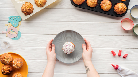 Cropped view of woman holding plate with cupcake on table with baking tools and traysの写真素材