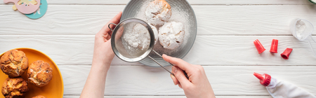 Panoramic shot of woman decorating cupcakes with powdered sugar on white table