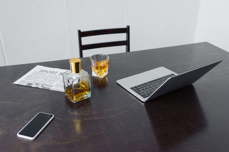 Wooden desk with laptop, sport news newspaper, smartphone with blank screen, bottle and glass of...