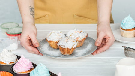 Cropped view of woman holding plate with cupcakes with powdered sugar on grey backgroundの写真素材