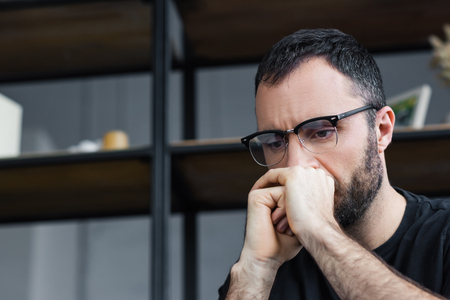 Thoughtful bearded man in glasses holding hands near face and looking down