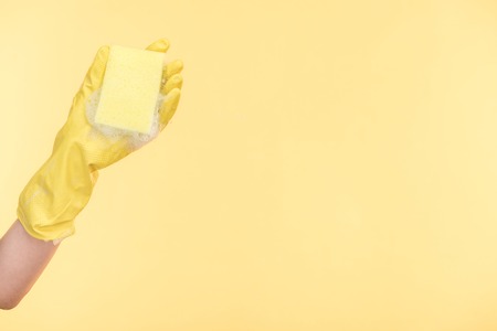 Cropped view of cleaner in yellow rubber glove holding sponge with bubbles on yellow background