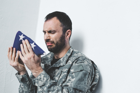 Depressed bearded military man crying while sitting by wall and holding USA national flagの写真素材