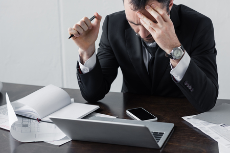 Thoughtful businessman sitting at workplace near laptop, documents and smartphone with blank screenの写真素材