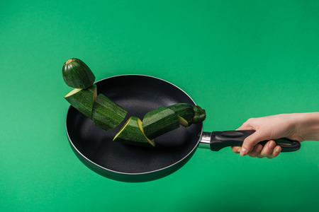 Partial view of woman holding frying pan with fresh green sliced zucchini on green backgroundの写真素材
