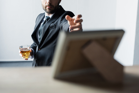 Selective focus of man in black suit holding glass of whiskey and outstretching hand to desk with photo frameの写真素材