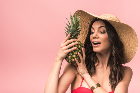 Amazed girl in straw hat and bikini holding pinapple isolated on pink backgroundの写真素材