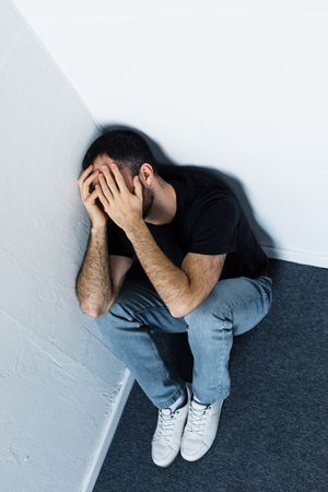High angle view of adult depressed man sitting on floor in corner and covering face with handsの写真素材